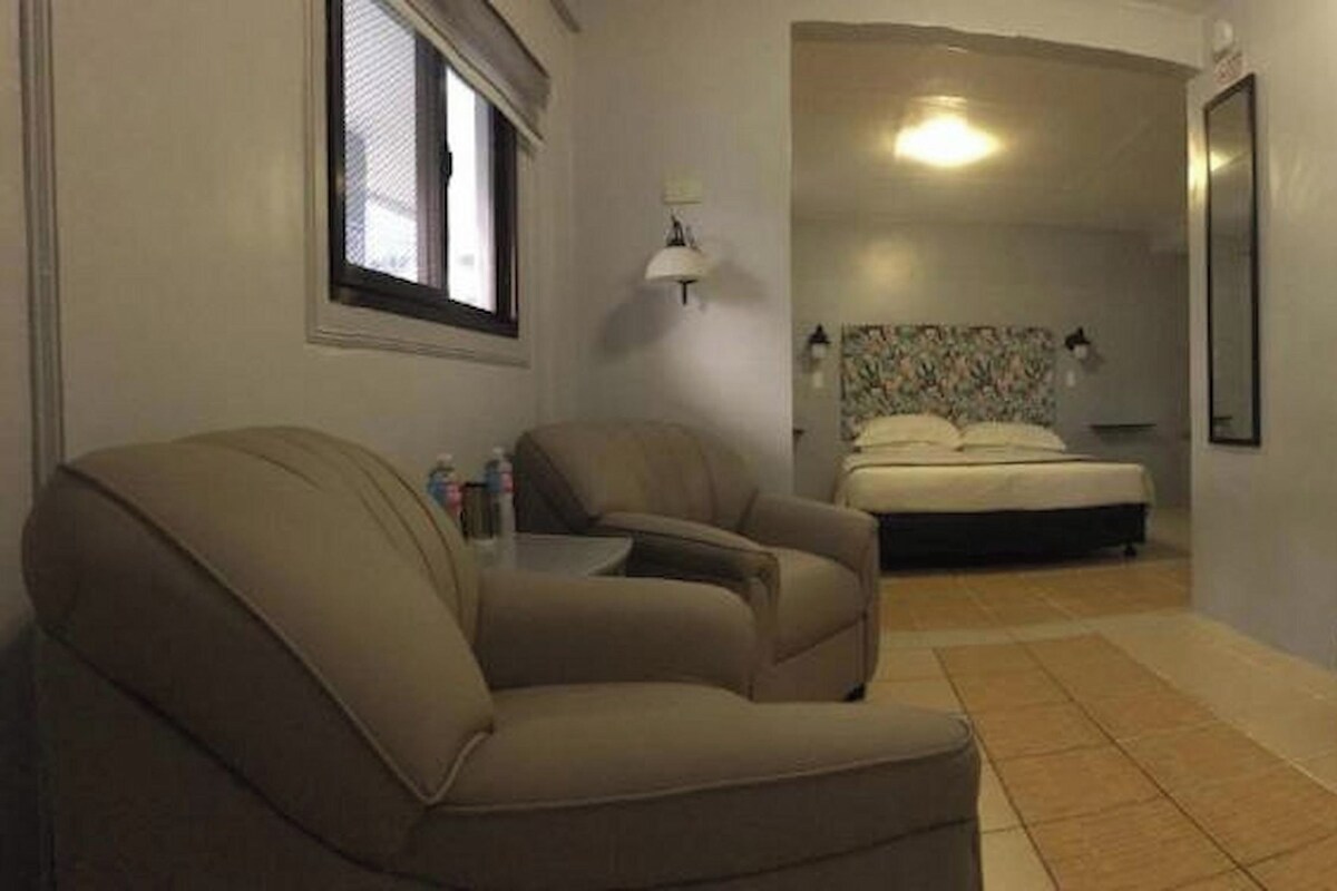Habitación pequeña y funcional con cama, una silla, una puerta de madera y un baño visible a través de una puerta abierta.