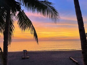 Escena de playa con palmeras, un banco y una pequeña estructura, presumiblemente un puesto de socorristas, bajo un cielo al atardecer.