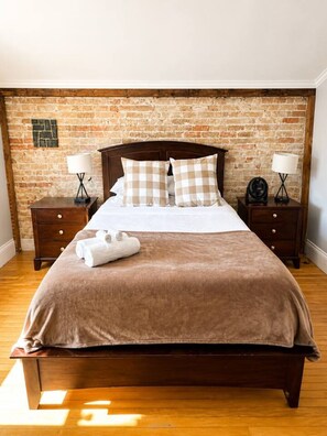 A bed with a brown blanket, two pillows, and a rolled towel on a wooden headboard. There are two bedside tables with lamps and a brick wall behind. - Private Pool Retreat | 6BR 3.5 BA | BBQ & Firepit (Cobourg)