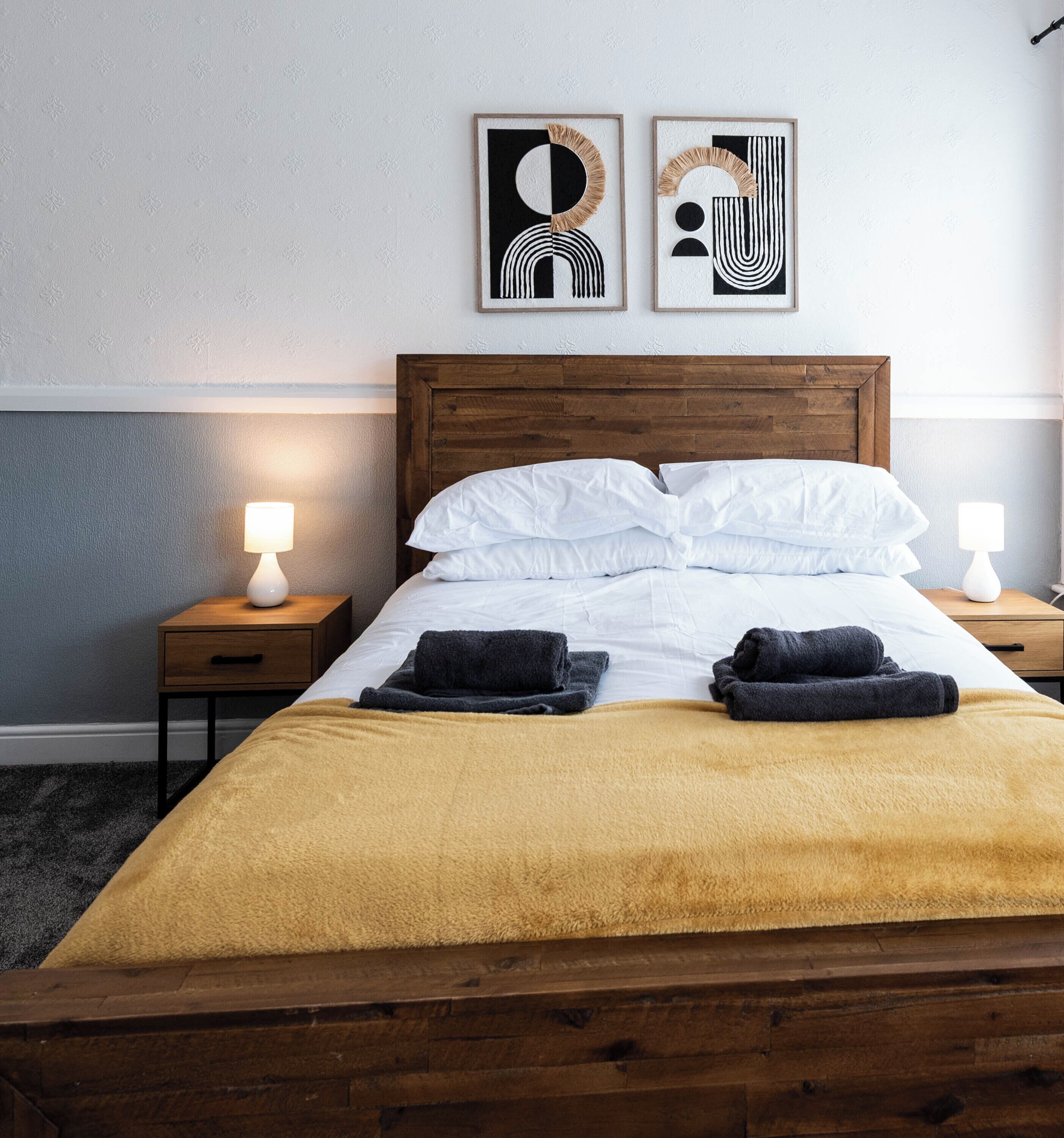 Un lit bien fait, avec du linge de lit blanc, un plaid jaune et deux serviettes noires. Il y a deux tables de chevet avec des lampes, et deux œuvres d'art abstraites accrochées au mur au-dessus du lit.