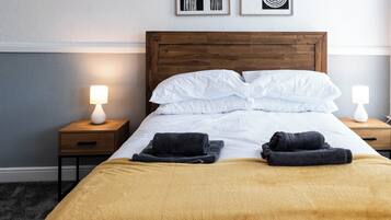 Un lit bien fait, avec du linge de lit blanc, un plaid jaune et deux serviettes noires. Il y a deux tables de chevet avec des lampes, et deux œuvres d'art abstraites accrochées au mur au-dessus du lit.
