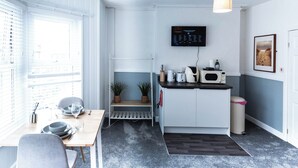 Eine kompakte Küche mit Mikrowelle, Toaster und Spüle, ein kleiner Esstisch mit Stühlen, ein an der Wand befestigter Fernseher und eine Tür zu einem weiteren Raum.