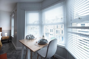 A dining area with a table set for two, a chair, and a window with blinds. - The Bay Apart-Hotel by Guestz (Morecambe)