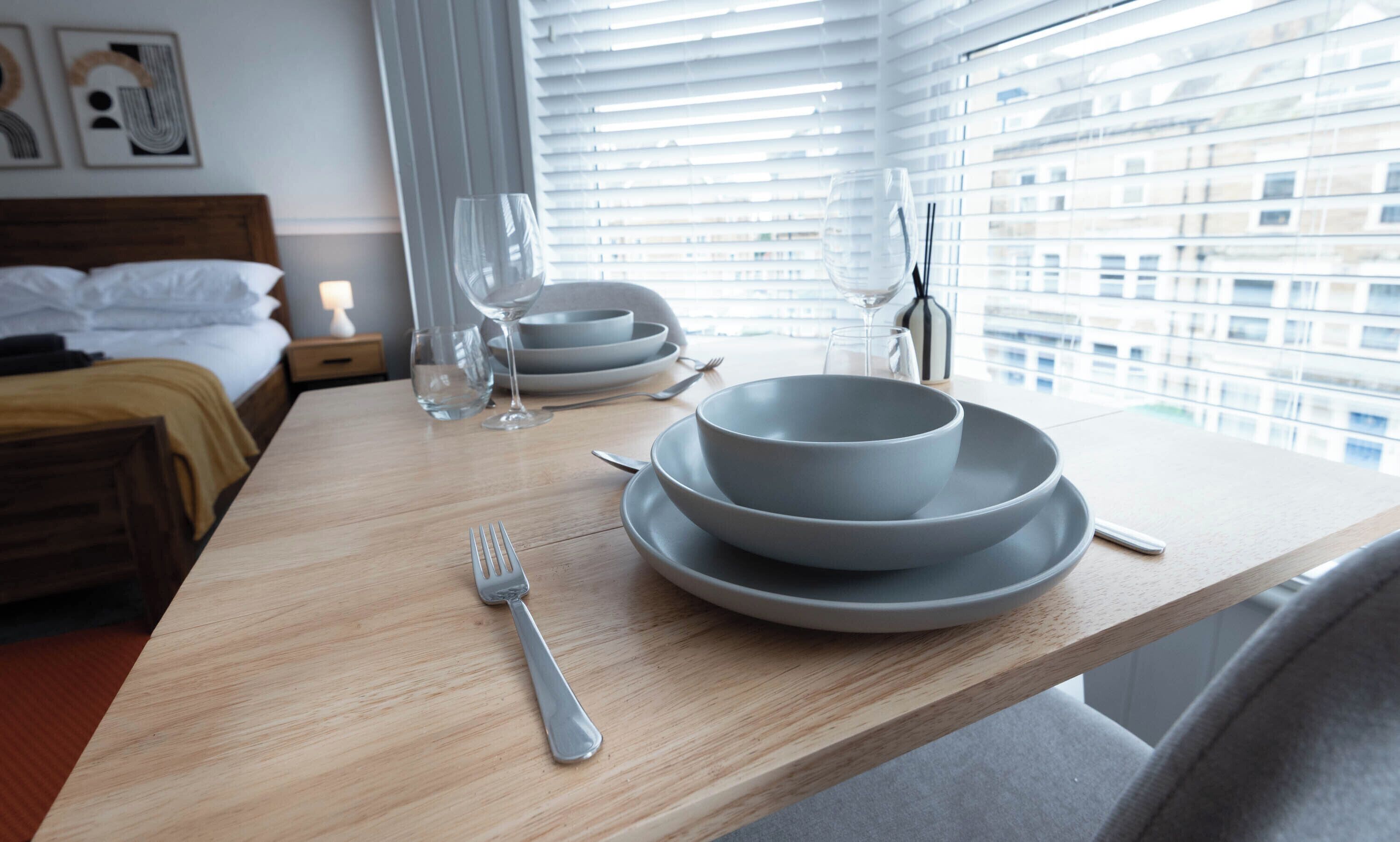 Una mesa de comedor bien dispuesta con platos, cuencos y cubiertos, una cama con cabecera de madera y una ventana con persianas.