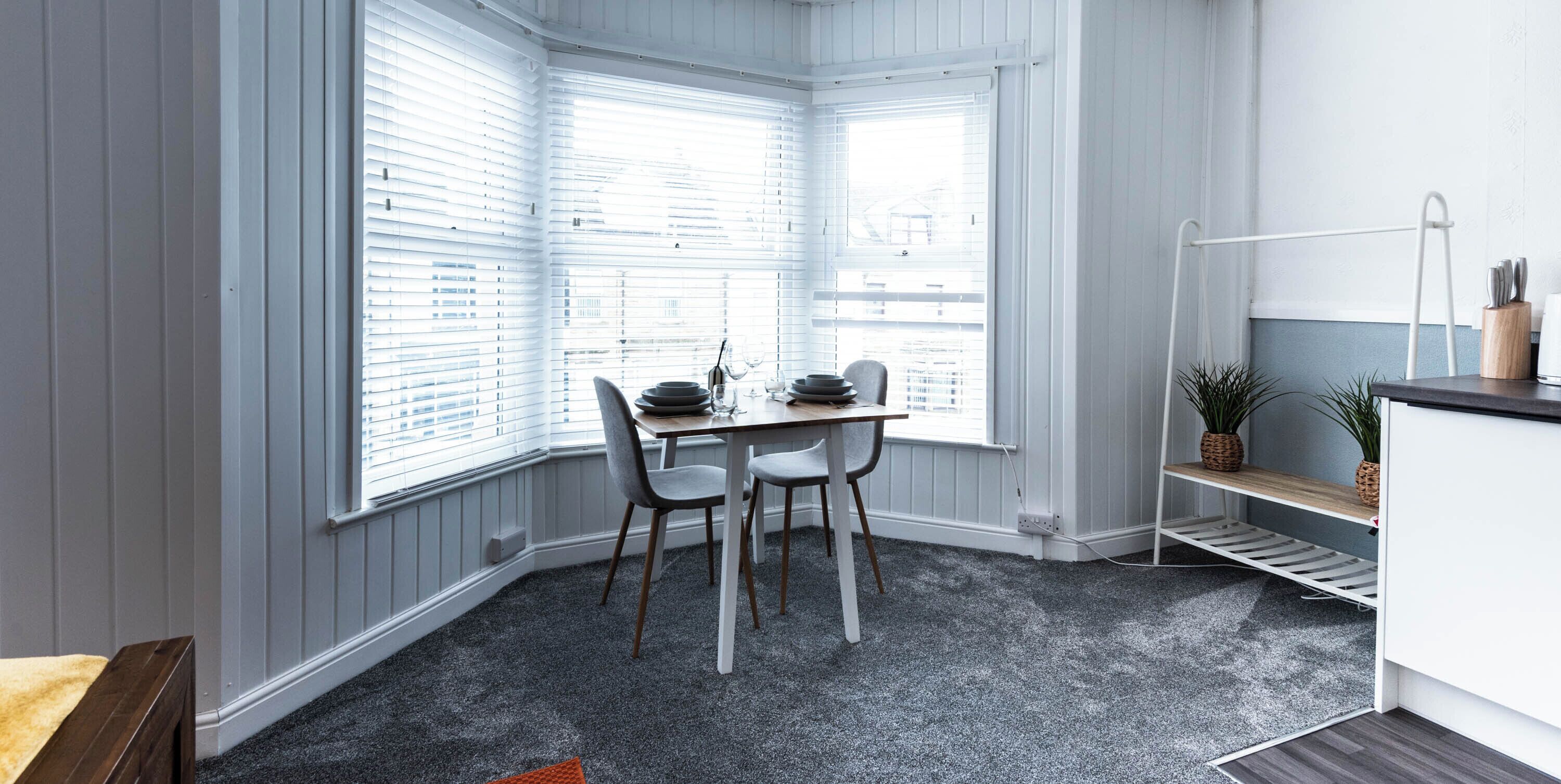 Habitación de esquina con mesa de comedor, dos sillas, una estantería pequeña con plantas y un radiador blanco.