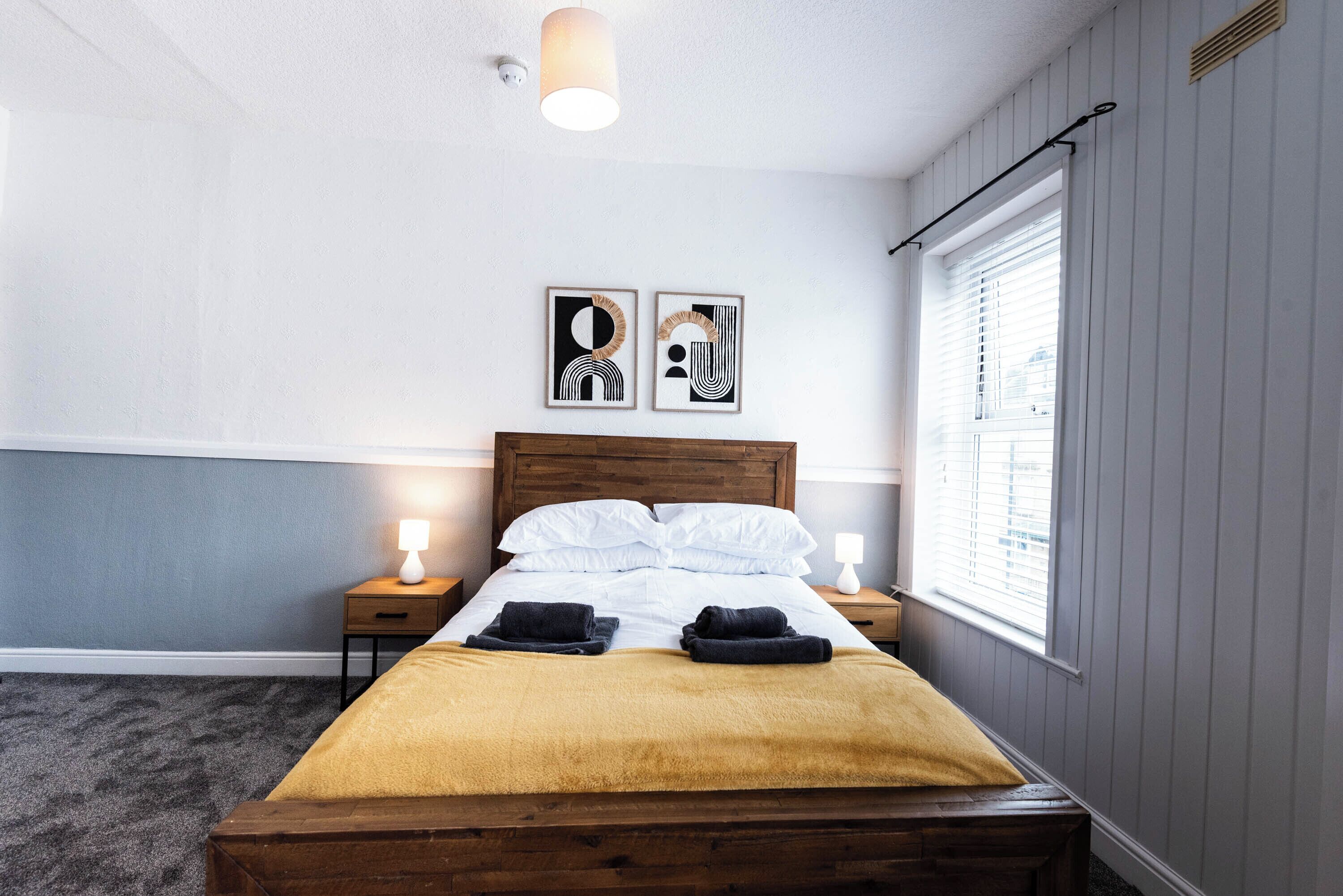A bedroom with a wooden bed, two bedside tables with lamps, a window with blinds, and two wall-mounted artworks.