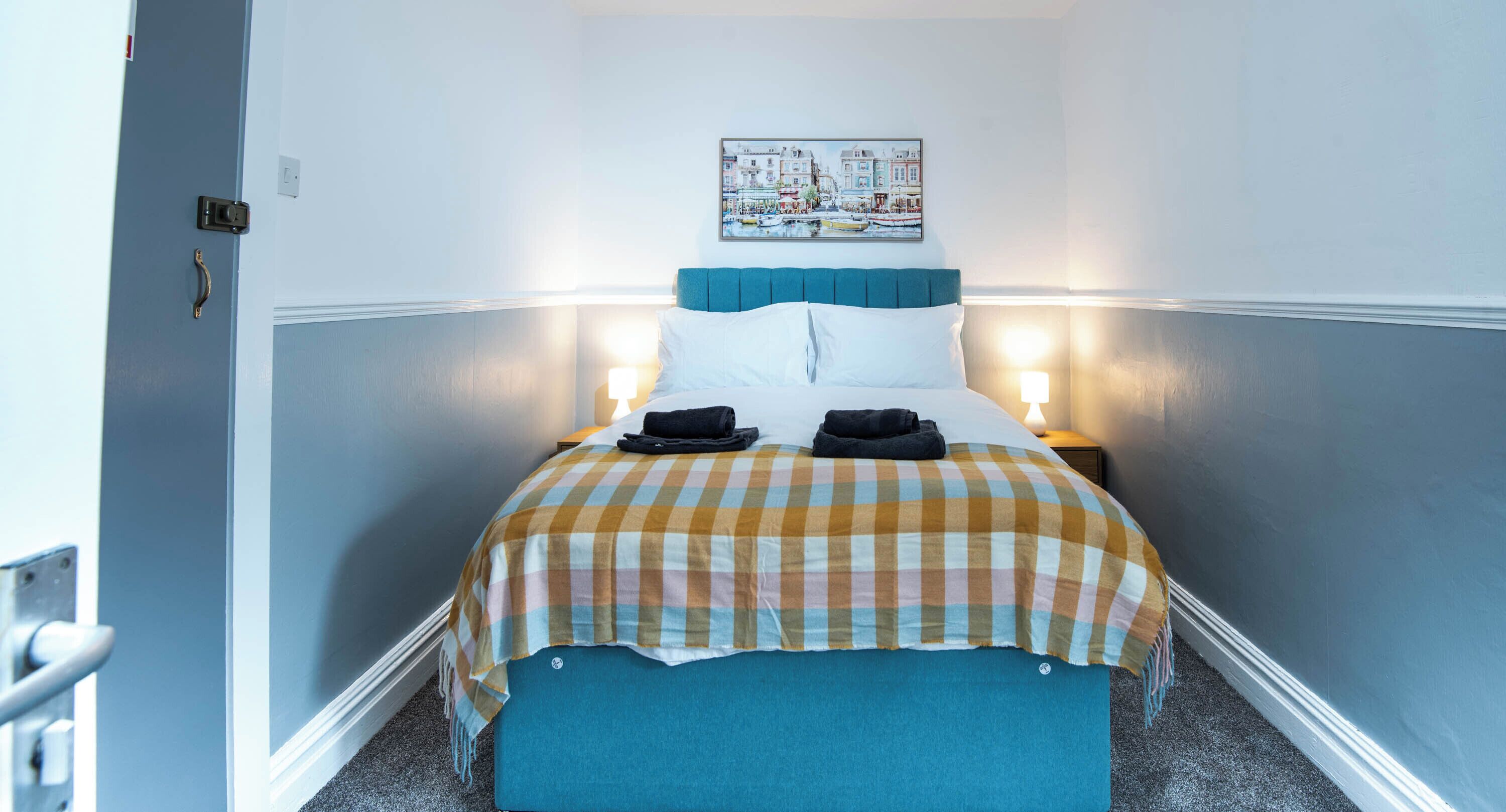 A hotel room with a bed, a checkered bedspread, a headboard, two bedside lamps, and a painting on the wall.
