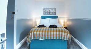 A hotel room with a bed, a checkered bedspread, a headboard, two bedside lamps, and a painting on the wall. - The Bay Apart-Hotel by Guestz (Morecambe)