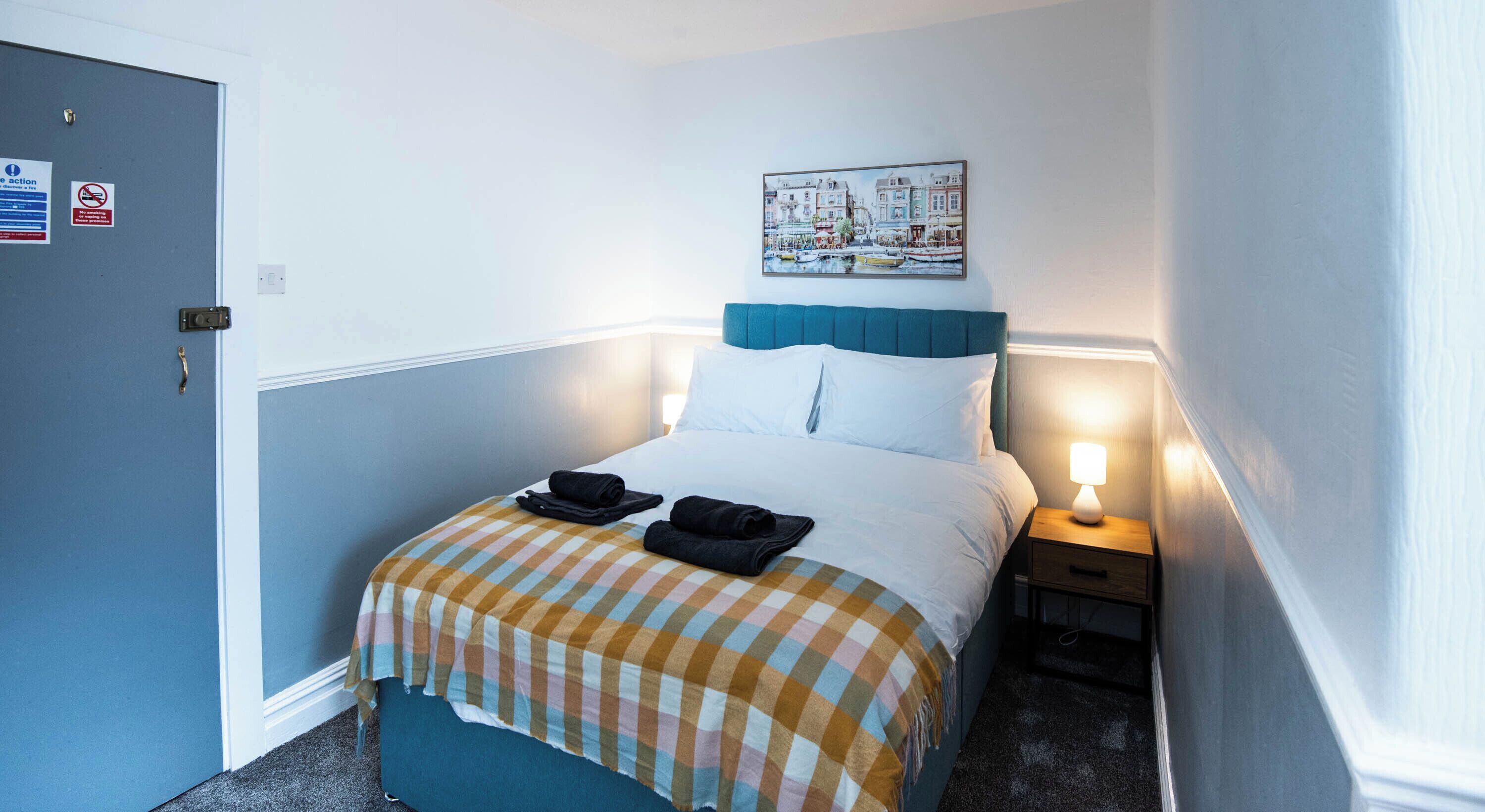 Una habitación de hotel con cama, mesita de noche, lámpara, un cuadro y una puerta.