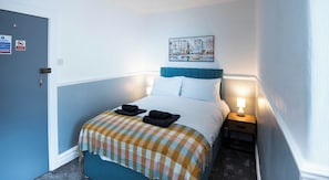 Habitació d'hotel amb un llit, una tauleta de nit, una làmpada, un quadre i una porta.