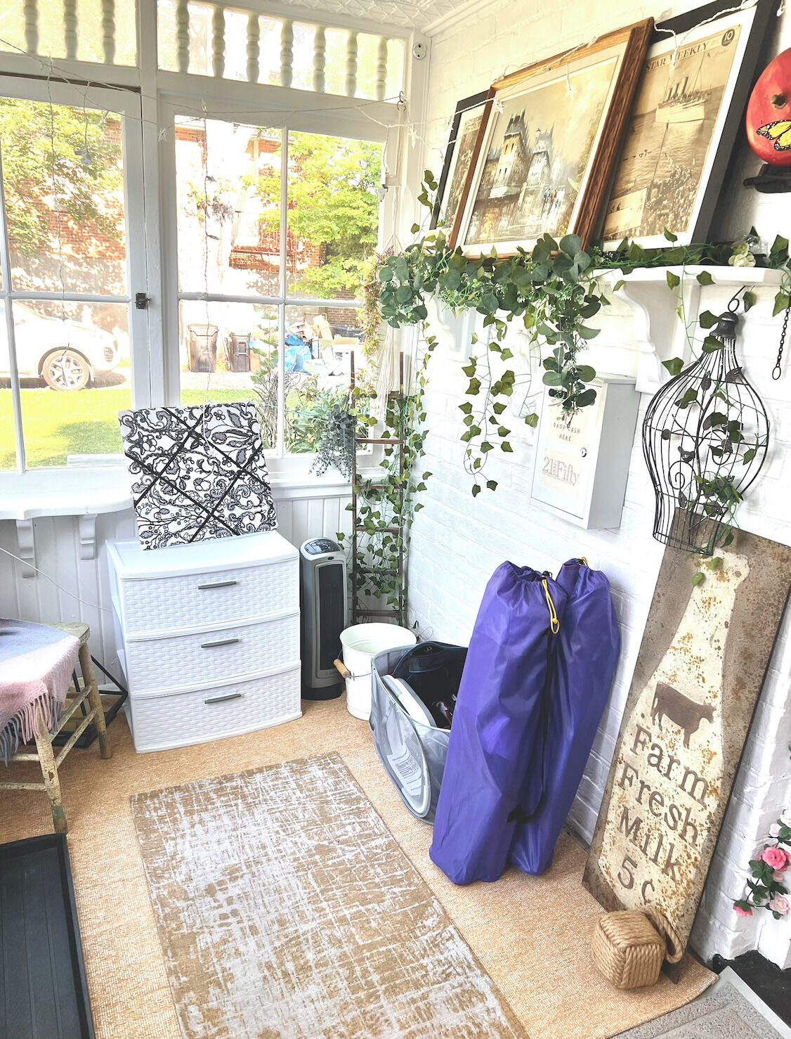 A room with a window, a white chest of drawers, a blue bag, a patterned pillow, and a wall with framed pictures and hanging plants.