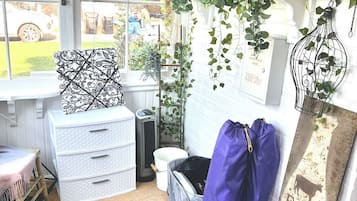 A room with a window, a white chest of drawers, a blue bag, a patterned pillow, and a wall with framed pictures and hanging plants.
