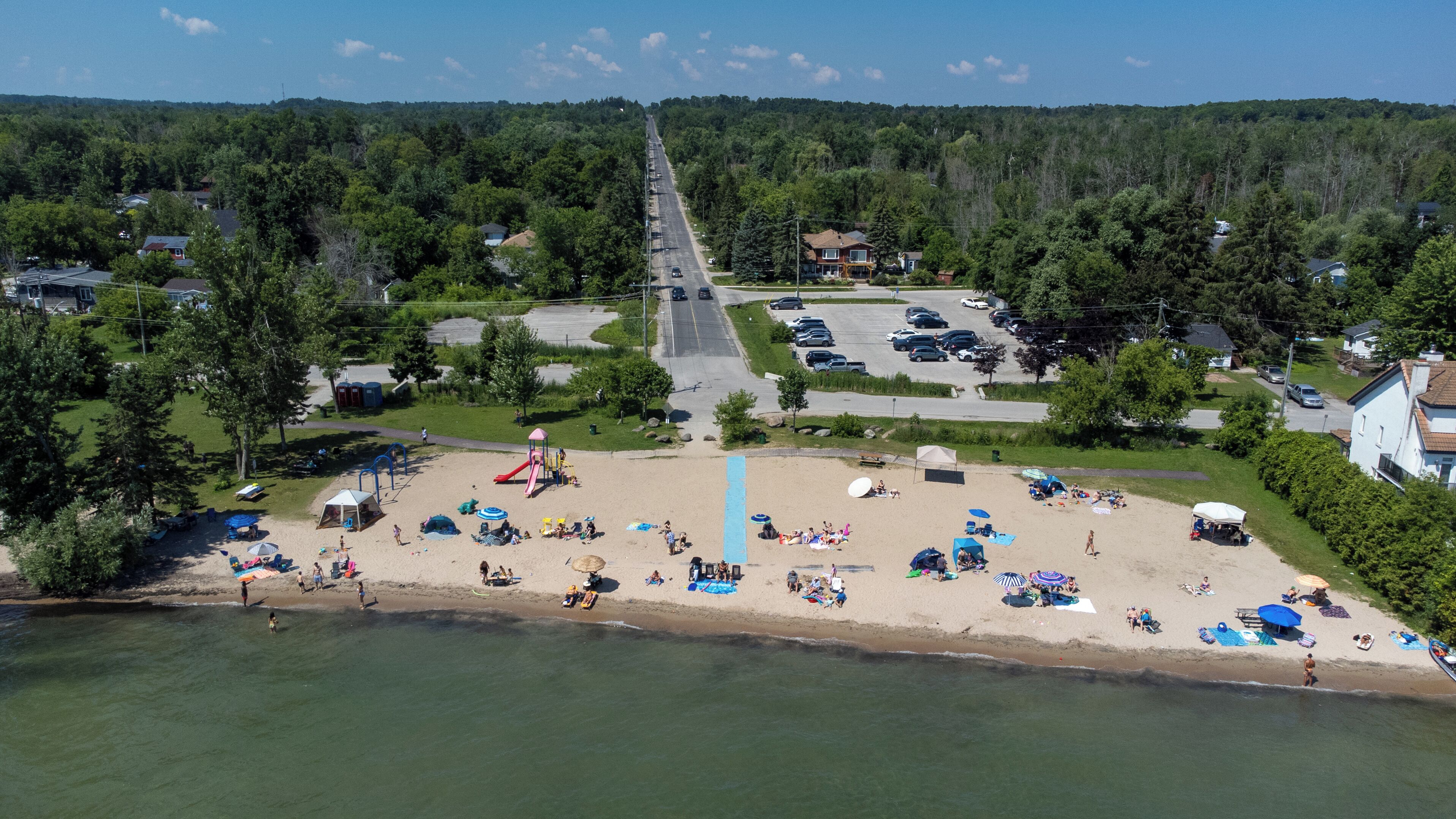 An aerial view of a beach with a playground, a parking lot, and residential houses in the background.