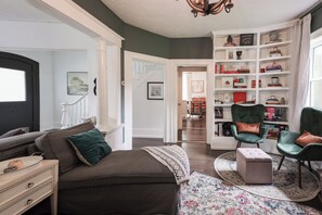 Ein Wohnzimmer mit einer Couch, einem Hocker und Sesseln, einem Bücherregal und einer Treppe.