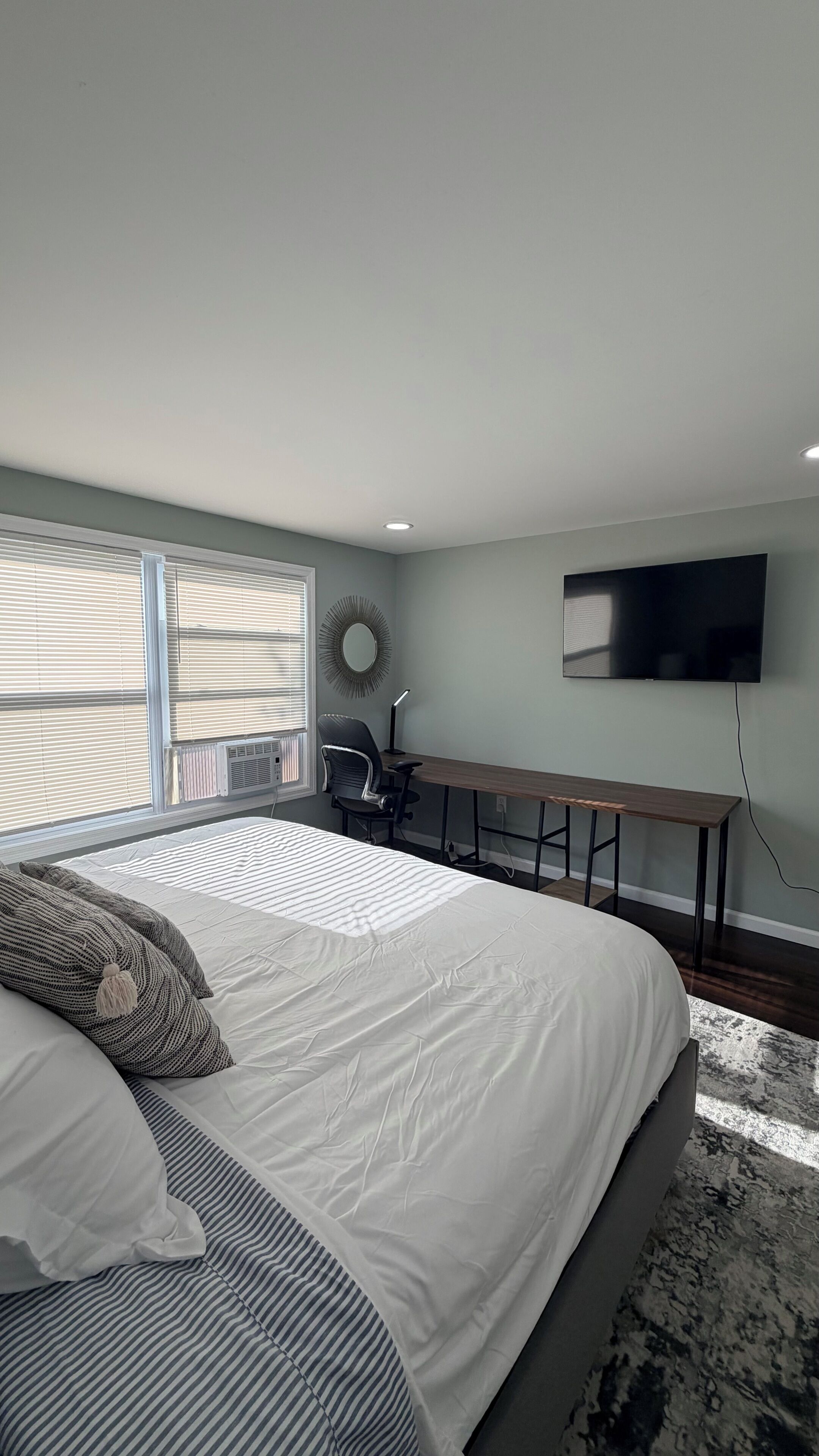 A bedroom with a bed, a desk, a chair, a TV, and a window with blinds.