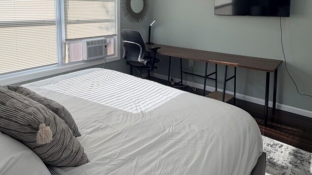 Ein Schlafzimmer mit Bett, Schreibtisch, Stuhl, Fernseher und einem Fenster mit Jalousien.