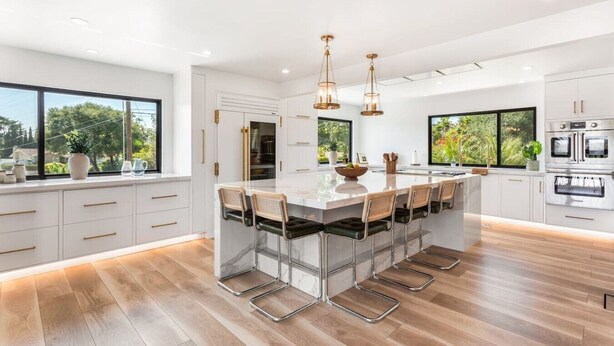 Una cocina moderna con una isla grande, taburetes de bar, electrodomésticos de acero inoxidable y ventanas amplias.