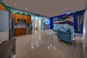 Ein modernes Wohnzimmer mit einem blauen Sofa, einer Küche mit Holzschränken, einem Fernseher und einem großen abstrakten Gemälde an der Wand.