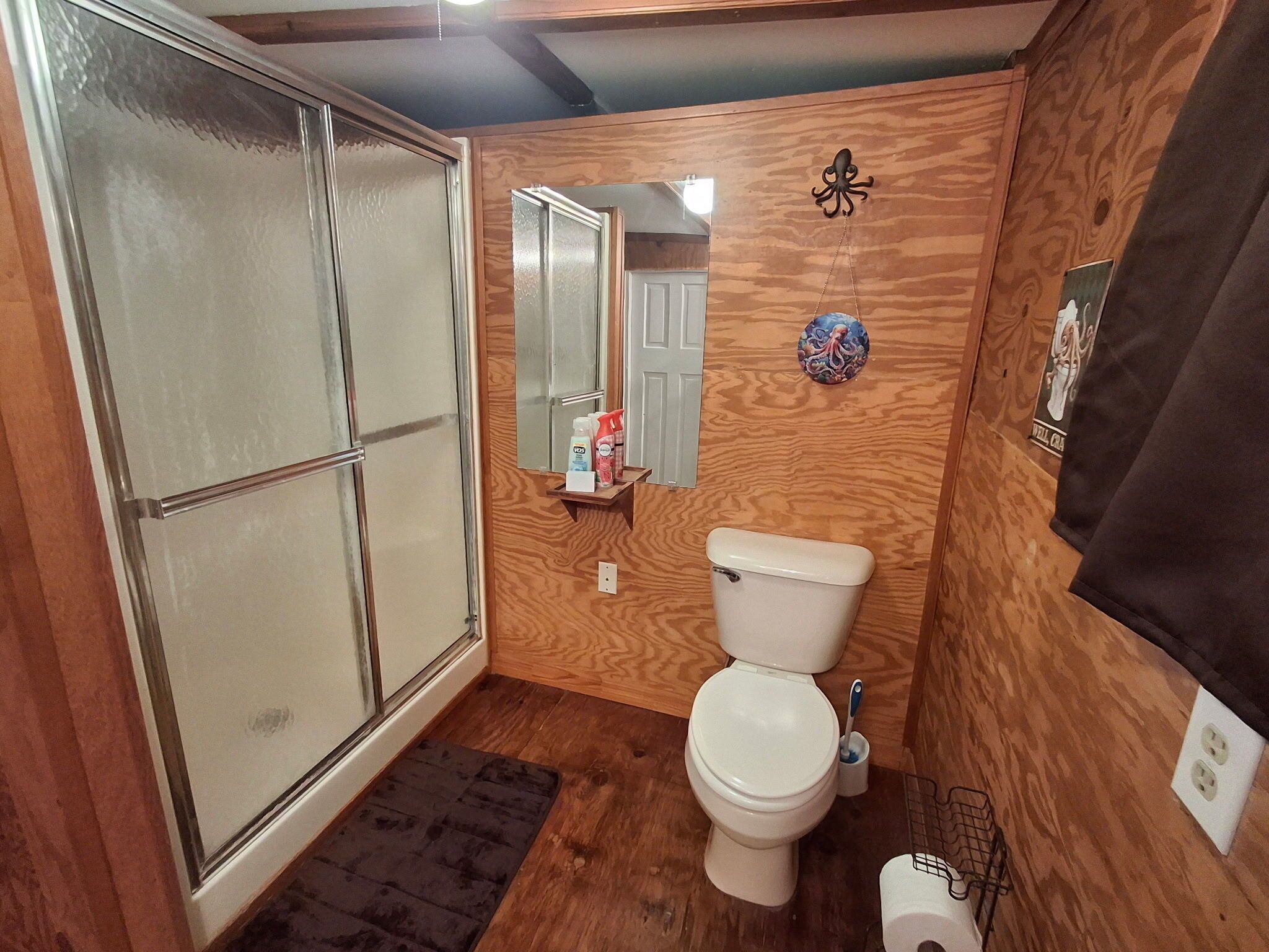 Un baño con pared de madera, un inodoro, una ducha con puertas de vidrio esmerilado y un pequeño estante con artículos de aseo.