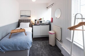 Quarto de hotel pequeno e funcional, com uma cama, uma escrivaninha com chaleira e torradeira, um radiador e uma janela com persianas.