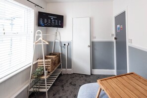 A hotel room with a TV mounted on the wall, a door, a window with blinds, a white chair, a wooden table, and a shelf with baskets. - The Bay Apart-Hotel by Guestz (Morecambe)