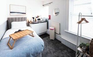 A compact hotel room with a bed, bedside table, a small kitchenette, a radiator, and a window with blinds. - The Bay Apart-Hotel by Guestz (Morecambe)