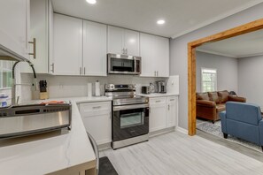 A modern kitchen with white cabinets, stainless steel appliances, and a wooden-framed doorway leading to a living area with a brown leather sofa and a blue armchair. - Delightful 3-bedroom house with AC in charming Decatur (Decatur)