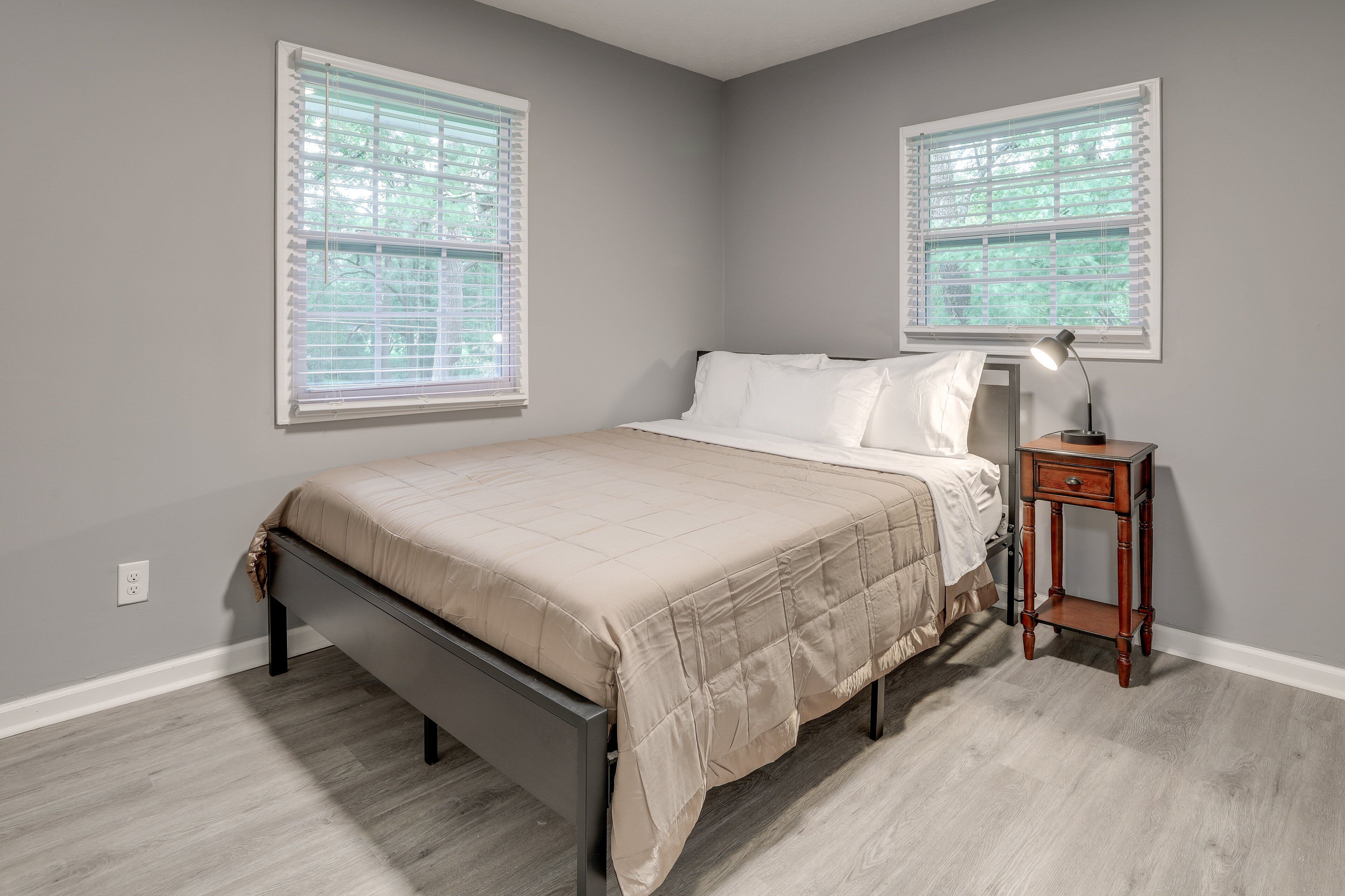 A bedroom with a bed, two windows, a bedside table with a lamp, and a power outlet on the wall.