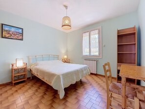A bedroom with a bed, bedside table, lamp, window, radiator, and a wooden chair. - L Escapade Orangeoise by Interhome (Orange)