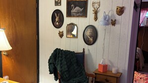 Ein Zimmer mit Holztür, einem Sessel mit Decke, einem kleinen Tisch mit Lampe sowie Wanddekorationen, darunter Tierköpfe und ein Schwanenbild.
