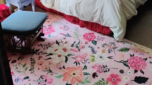 Ein Schlafzimmer mit einem Bett, einem Nachttisch, einem Sessel, einem Spiegel und einem Fenster mit karierten Vorhängen.