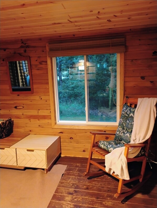 Ein Holzhütten-Innenraum mit einem Schaukelstuhl, einem kleinen Tisch und einem Fenster mit Blick auf einen Wald.