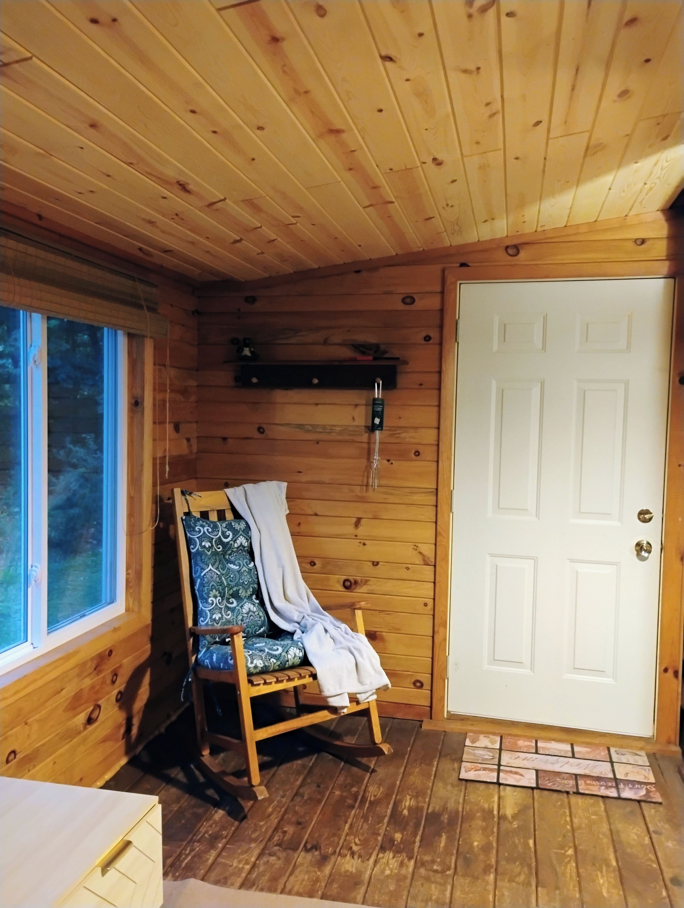 Interior de uma cabana de madeira, com uma cadeira de balanço, uma porta branca e uma janela.