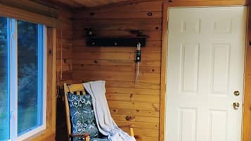 Interior de uma cabana de madeira, com uma cadeira de balanço, uma porta branca e uma janela.