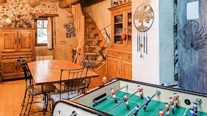 Une pièce chaleureuse avec une table à manger en bois, des chaises, un baby-foot, un escalier et un mur décoré de différents objets.