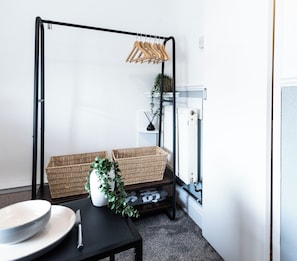 A room with a black metal stand holding wooden hangers, a wicker basket, a white vase with green foliage, and a black table with a white bowl and a knife. - The Bay Apart-Hotel by Guestz (Morecambe)
