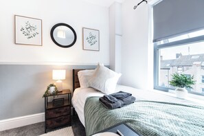 A bedroom with a bed, a nightstand, a bedside lamp, a window with a view, and framed artwork on the wall. - The Bay Apart-Hotel by Guestz (Morecambe)