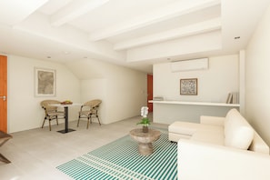 A modern living room with a white sofa, a dining table with wicker chairs, a small round table with a vase of flowers, and a patterned rug. - Kandooma Maldives - All Inclusive with Kids Stay & Eat Free (Kandooma)