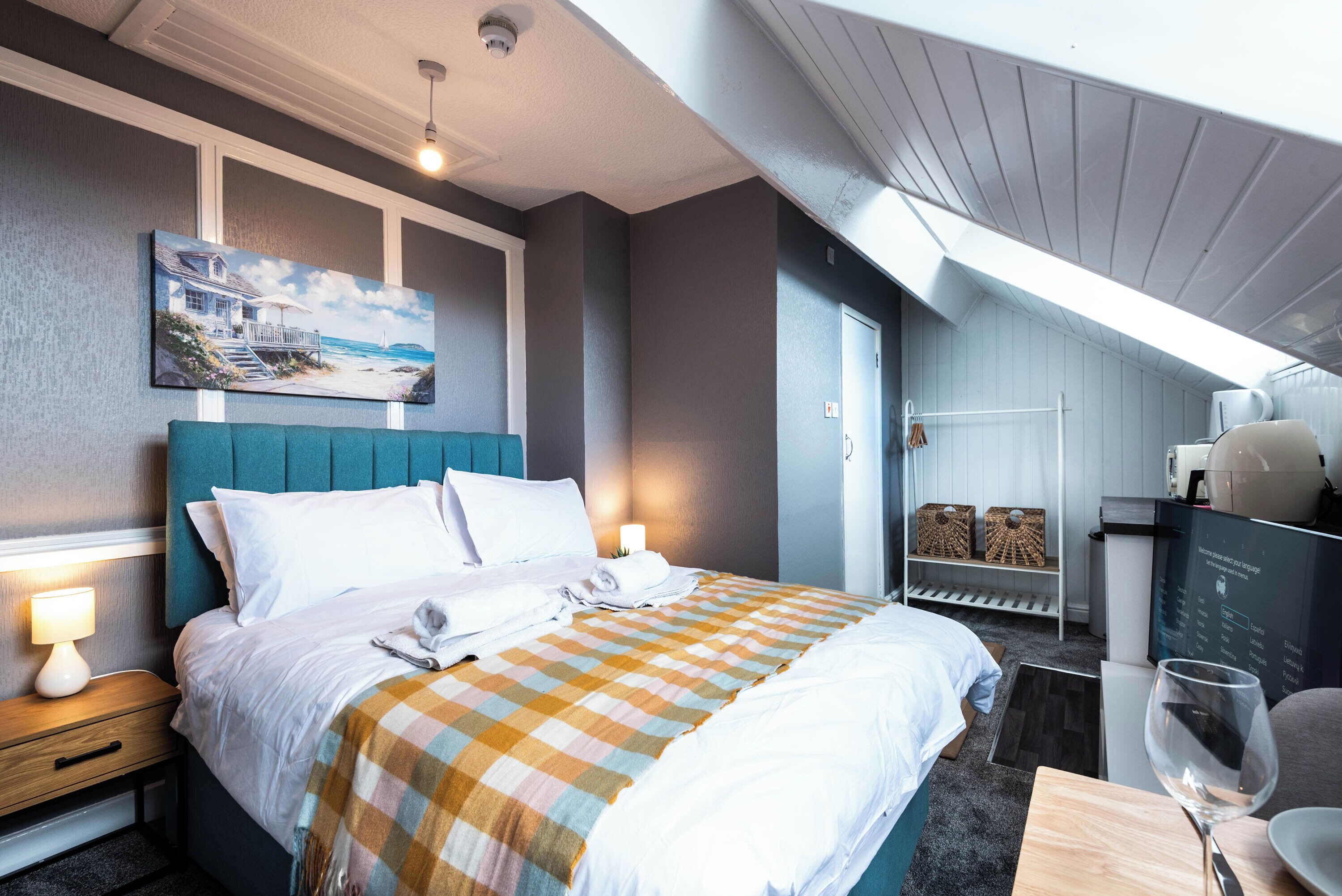 A hotel room with a bed, bedside lamps, a painting, a checkered bedspread, a desk with a glass, and a coffee machine.