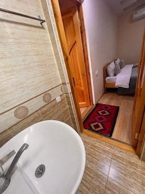A bathroom with a sink, a shower, and a doorway leading to a bedroom with a bed and a red rug. - EmirA (Samarkand)