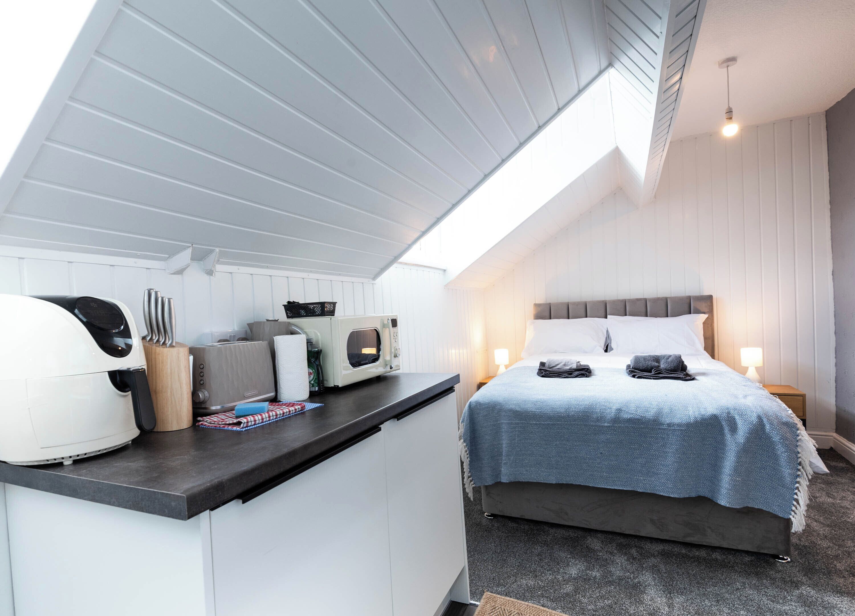Una habitación pequeña con kitchenette, una cama con una manta azul y una mesita de noche con una lámpara.