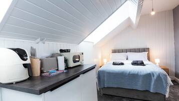 Una habitación pequeña con kitchenette, una cama con una manta azul y una mesita de noche con una lámpara.