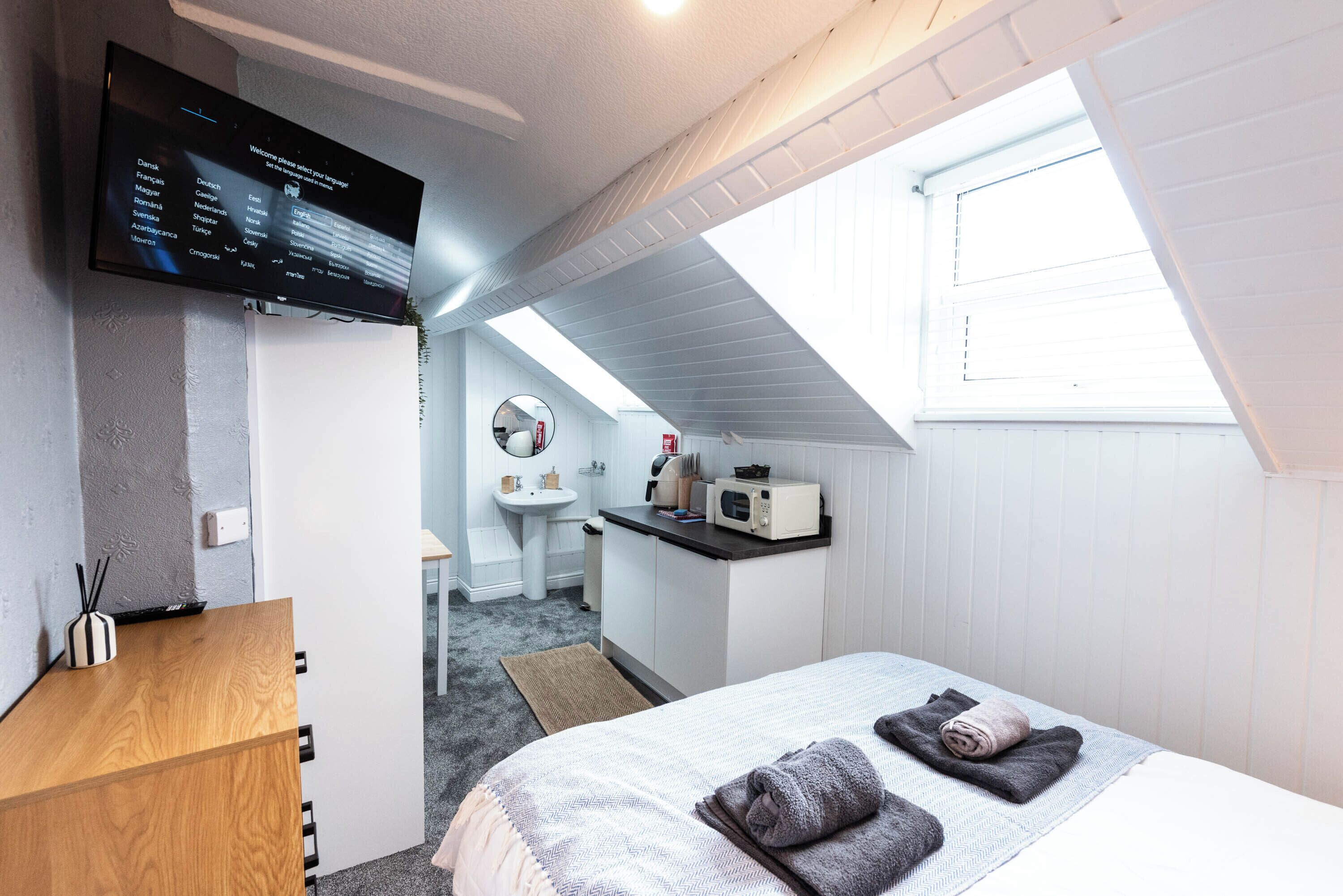 A bedroom with a TV, a desk, a bed with towels, a sink, and a mirror.
