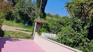 A pink outdoor area with steps leading to a covered space, surrounded by greenery and a clear blue sky. - The Rainbow House (Roma)
