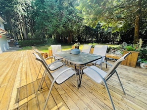 Una terraza al aire libre de madera con una mesa de vidrio y sillas de metal, rodeada de follaje.