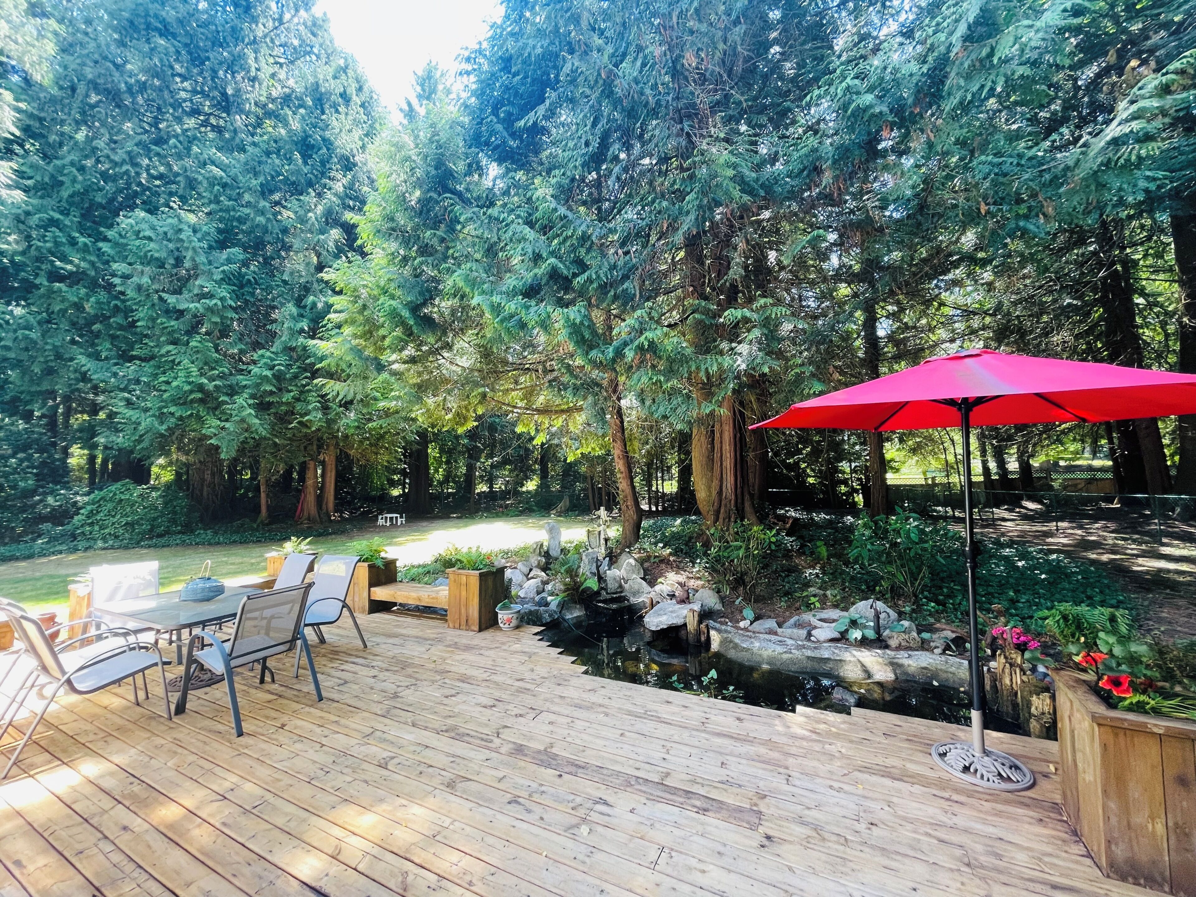 Una terraza de madera con mobiliario exterior, un paraguas rojo y un estanque con rocas y plantas.