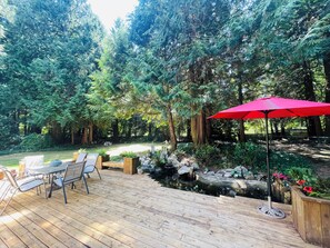 Una terraza de madera con mobiliario exterior, un paraguas rojo y un estanque con rocas y plantas.