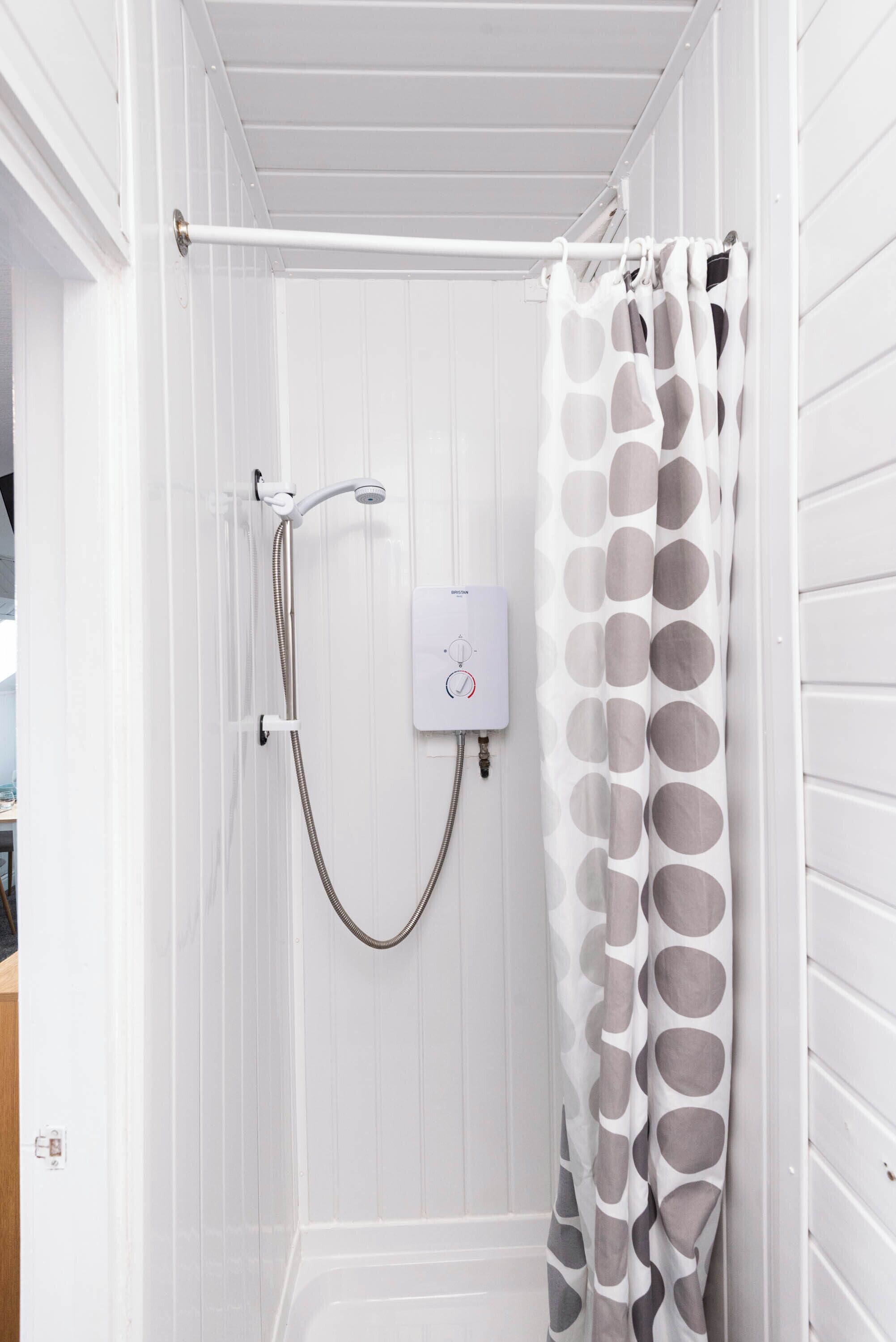 A small, white shower room with a showerhead and control panel, a towel rack, and a polka-dotted shower curtain.