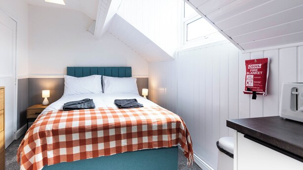 Ein Schlafzimmer mit karierter Bettdecke, einem türkisblauen Kopfteil, Nachttischlampen, einer roten Feuerschutzdecke an der Wand und einem Fenster über dem Bett.