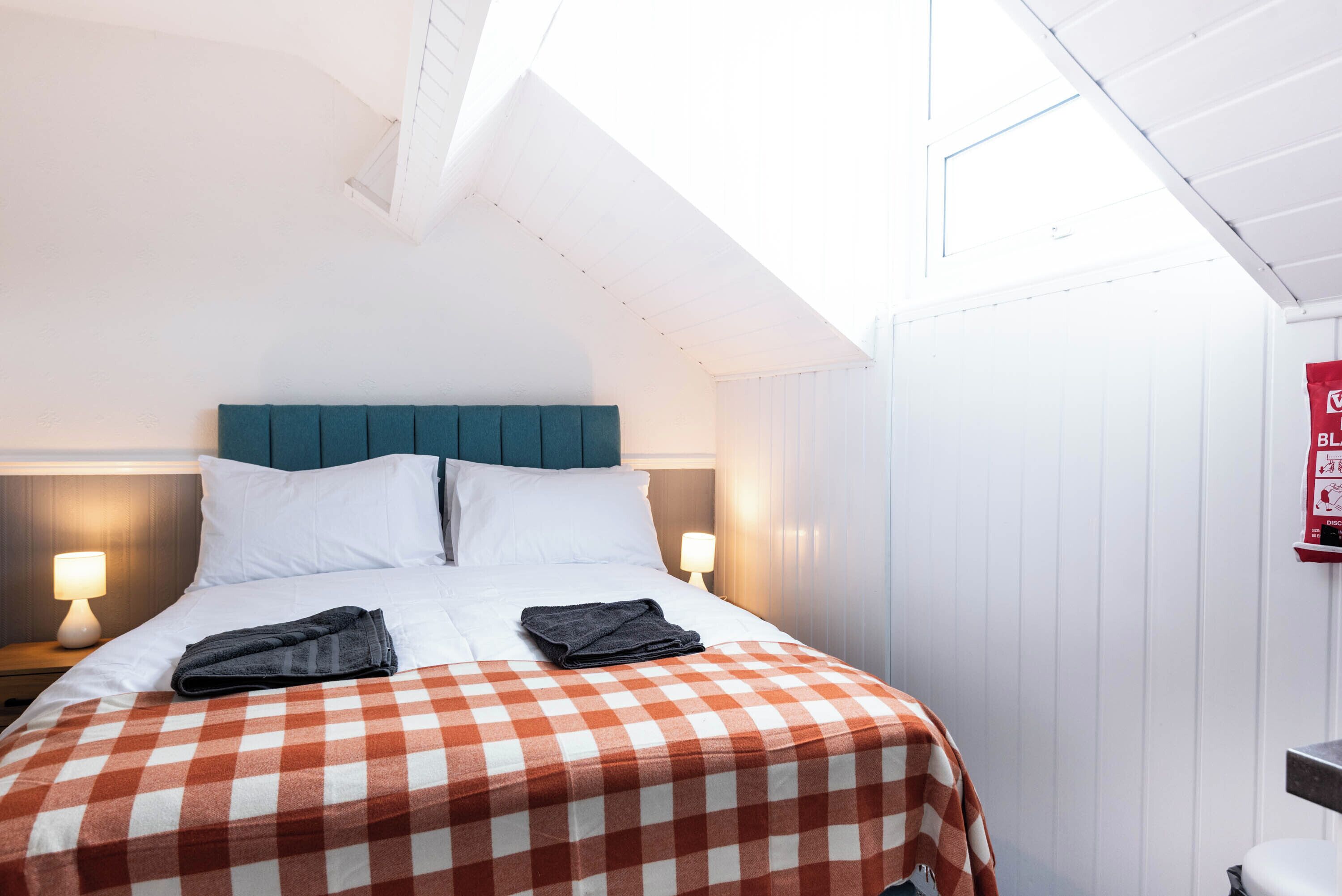 A hotel room with a bed, checkered bedspread, two pillows, a nightstand with a lamp, and a window with a view of the sky.